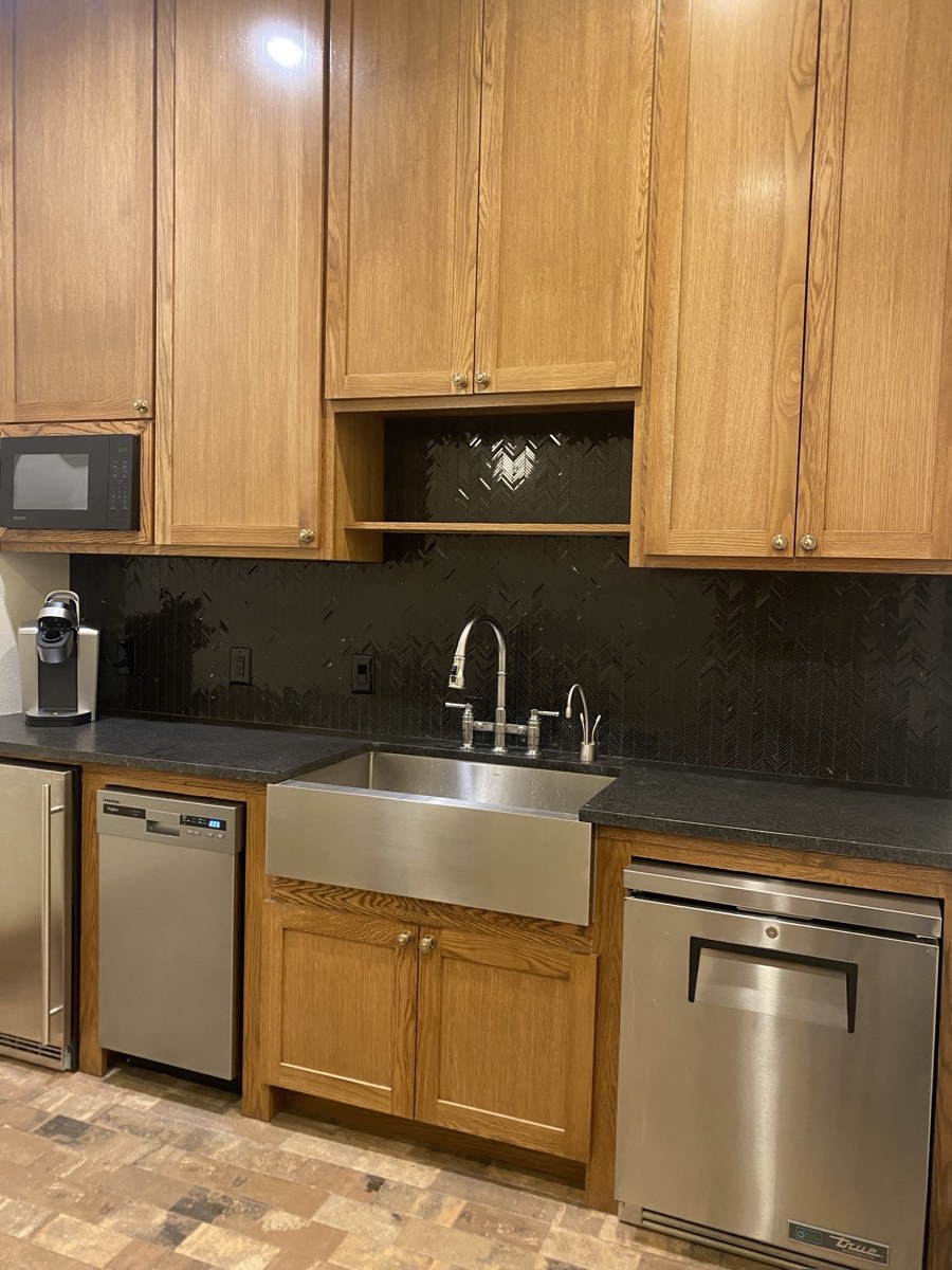 Kitchen with warm oak cabinetry, a black herringbone tile backsplash, and a stainless steel farmhouse sink